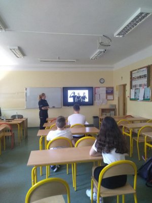 zdjęcie przedstawia salę lekcyjną w której znajduje się policjantka przeprowadzająca zajęcia profilaktyczne oraz dzieci siedzące w ławkach, na tablicy wyświetlany jest spot Narkotyki i dopalacze zabijają, szkoda Ciebie na patoklimaty