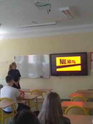 zdjęcie przedstawia salę lekcyjną w której znajduje się policjantka przeprowadzająca zajęcia profilaktyczne oraz dzieci siedzące w ławkach, na tablicy wyświetlany jest spot Narkotyki i dopalacze zabijają, szkoda Ciebie na patoklimaty