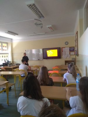 zdjęcie przedstawia salę lekcyjną w której znajduje się policjantka przeprowadzająca zajęcia profilaktyczne oraz dzieci siedzące w ławkach, na tablicy wyświetlany jest spot Narkotyki i dopalacze zabijają, szkoda Ciebie na patoklimaty