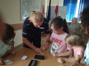 zdjęcie wykonane jest w pomieszczeniu, widać na nim policjantkę pochyloną nad biurkiem i wykonującą odbitkę linii papilarnych dziewczynce, w pomieszczeniu znajduje się kilkoro dzieci, za policjantką znajduje się stojak z trzema flagami, tablica oraz mapa