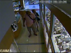 Zdjęcie przedstawia wnętrze sklepu z biżuterią, w którym znajdują się trzy osoby. Mężczyzna  o krępej budowie ciała ubrany jest w koszulkę z krótkim rękawem w jasnym kolorze, jasne spodenki długością sięgające do kolan, klapki typu japonki. Na głowie ma jasną czapkę z daszkiem, przez ramię przewieszoną torbę w ciemnym kolorze. Kobieta stojąca za nim ma ciemne upięte włosy, ubrana jest w jasne długie spodnie, jasną bluzkę, ciemne nakrycie wierzchnie, na lewym ramieniu ma przewieszoną ciemną torebkę. Druga kobieta stoi w pobliżu wyjścia, ubrana jest w jasną bluzkę z krótkim rękawem, ma długie ciemne rozpuszczone włosy.