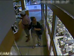 Zdjęcie przedstawia wnętrze sklepu z biżuterią, w którym znajdują się trzy osoby. Mężczyzna  o krępej budowie ciała ubrany jest w koszulkę z krótkim rękawem w jasnym kolorze, jasne spodenki długością sięgające do kolan, klapki typu japonki. Na głowie ma jasną czapkę z daszkiem, przez ramię przewieszoną torbę w ciemnym kolorze. Mężczyzna w wyciągniętą rękę w stronę jednej z gablot. Kobieta stojąca za nim ma ciemne upięte włosy, ubrana jest w jasne długie spodnie, jasną bluzkę, ciemne nakrycie wierzchnie, na lewym ramieniu ma przewieszoną ciemną torebkę. Druga kobieta stoi w pobliżu wyjścia, ubrana jest w jasną bluzkę z krótkim rękawem, ma długie ciemne rozpuszczone włosy.
