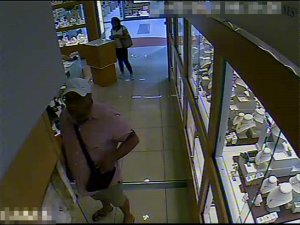 Zdjęcie przedstawia wnętrze sklepu z biżuterią, w którym znajdują się dwie osoby. Mężczyzna  o krępej budowie ciała ubrany jest w koszulkę z krótkim rękawem w jasnym kolorze, jasne spodenki długością sięgające do kolan, klapki typu japonki. Na głowie ma jasną czapkę z daszkiem, przez ramię przewieszoną torbę w ciemnym kolorze. Patrzy w stronę kamery. Kobieta, która stoi niedaleko wejścia do sklepu ubrana jest w jasną bluzkę z krótkim rękawem, długie ciemne spodnie, buty sportowe, ma długie ciemne rozpuszczone włosy. Przez lewe ramię ma przewieszoną torebkę.