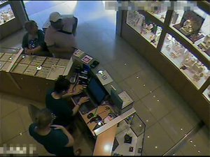 Zdjęcie przedstawia wnętrze sklepu z biżuterią. Przy komputerze, za ladą znajdują się pracownice sklepu, obie kobiety mają na sobie ciemne ubrania. Jedna stoi a druga siedzi na krześle. Po drugiej stronie lady stoją dwie osoby, mężczyzna i kobieta. Mężczyzna ubrany jest w czapkę z daszkiem, jasną koszulkę, lewym przedramieniem oparty jest o gablotę. Przy tej samej gablocie stoi kobieta, ubrana z bluzkę z nadrukiem w kształcie serca, w ciemne wierzchnie okrycie, trzyma ręce spleciona na gablocie, głowę ma zwróconą w lewą stronę.